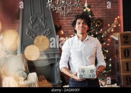 Ritratto di Allegro dai capelli ricci guy. Giovane uomo in piedi nella bella sala decorata e azienda scatola regalo bianca nel nuovo anno di tempo Foto Stock