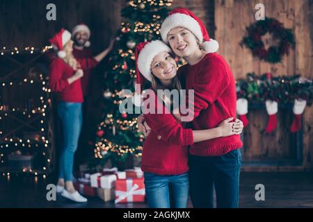 Foto di papà, mamma e due bambini aventi migliori X-mas eve insieme un ragazzo e una ragazza abbracciando vicino a garland tree usura interni santa cappucci e maglioni rosso Foto Stock