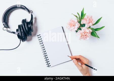 La donna è la scrittura a mano in Blocco note vuote nei pressi di cuffie e bouquet di fiori sulla scrivania bianca, piatto laici. Area di lavoro femminile nozione. Foto Stock