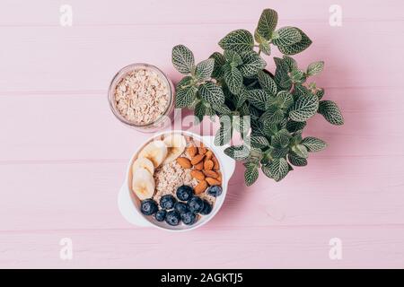 Sana colazione fiocchi d'avena con mirtilli, mandorle e banane accanto al vaso e pianta verde sulla tabella di rosa, vista dall'alto. Foto Stock