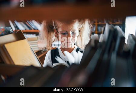 Bambina con gli occhiali alla ricerca di un libro in biblioteca. Concezione dell'istruzione Foto Stock