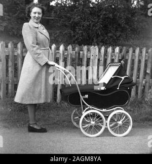 Anni '1950, storica, una signora con un cappotto in piedi all'esterno che tiene il manico di una carrozza per bambini o carrozza tradizionale a quattro ruote dell'epoca, Inghilterra, Regno Unito. Foto Stock