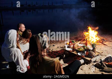 Amici in appoggio sulla spiaggia la sera, sono seduta dal fuoco e mangiare frutta fresca e frutti di mare Foto Stock