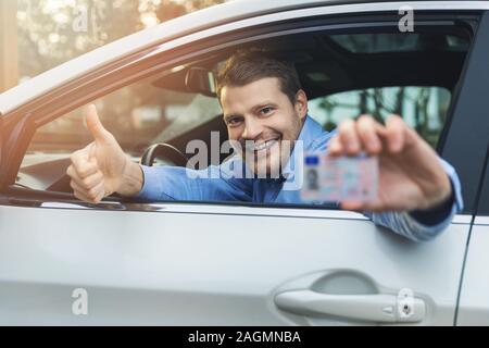 Giovane uomo sorridente seduto in macchina e che mostra la sua nuova licenza di pilota con il pollice in alto segno al di fuori della finestra di auto Foto Stock
