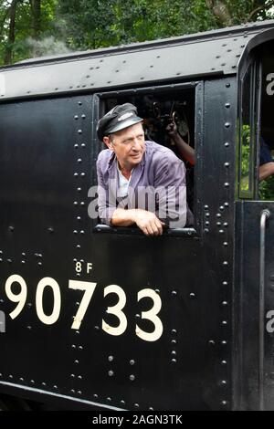 Il conducente della locomotiva a vapore 8F 2-8-0 numero 90733 si appoggia al di fuori della cabina mentre il motore è in piedi sulla stazione Oxenhope vicino a Keighley, Yorkshire. Foto Stock