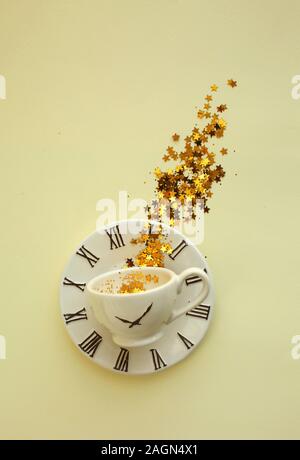 Una tazza di caffè con tinsel e decorazione di stelle dorate all'interno, posto sulla piastra di piccole dimensioni con orologio icona. Sfondo giallo. Foto Stock