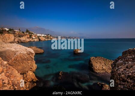 Sezione di spiaggia sulla costa spagnola di Costa del Sol in Nerja. Giornata soleggiata con blu delle acque del Mediterraneo con rocce e città in background Foto Stock