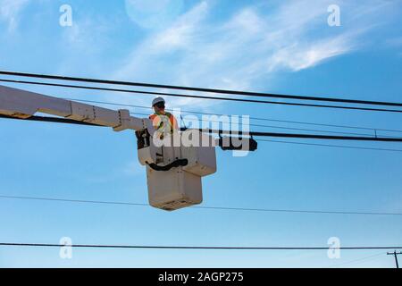 Un basso angolo di visione di un servizio di utilità infrastructure engineer lavorando in una piattaforma di lavoro aerea contro un cielo blu con nuvole wispy Foto Stock