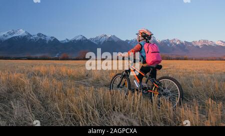 Uno dei bambini caucasici a piedi con bici nel campo di grano. Poco ragazza camminare nero ciclo arancione su sfondo di belle montagne innevate. Foto Stock