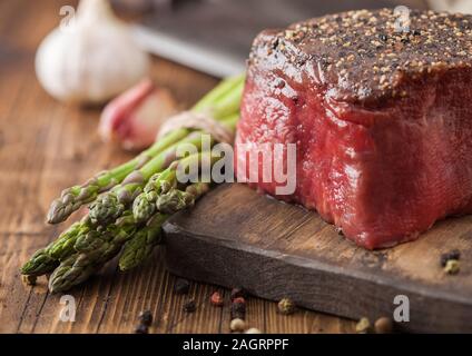 Fetta di manzo crudo fesa giunto con il sale e il pepe sul tagliere di legno con aglio e punte di asparagi su legno tavolo da cucina. Foto Stock