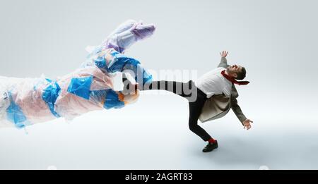 Giovane uomo caucasico evitando grandi mano in plastica bianca su sfondo per studio. Modello maschile in ufficio wear combattimenti in movimento e di azione. Guarda arrabbiato, spaventata, spinti. Problemi di eco, inquinamento, riciclaggio. Foto Stock