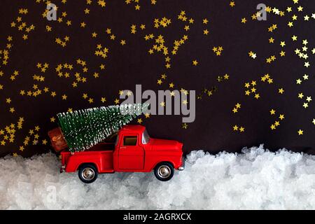 Rosso miniaturizzati giocattolo auto porta un albero di Natale sul tetto nella neve su sfondo scuro con stelle luccicanti con copia spazio. Composizione di natale Foto Stock