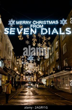 Le luci di Natale al di sopra della strada dicendo Buon Natale da Seven Dials con il simbolo del fiocco di neve con decorazioni in background, sera, Londra Centrale. Foto Stock