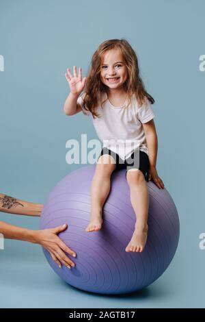 Ragazza seduta su un lilla sfera fitness e agitando la mano. Foto Stock