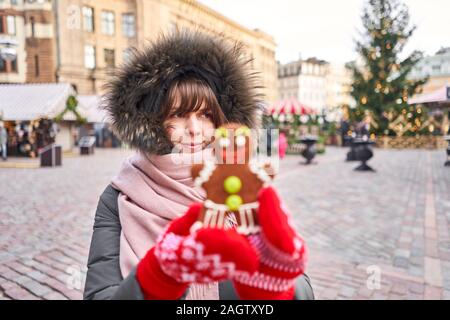 Mano nella Red mitten tenendo un sorridente omino di pan di zenzero e Natale umore a sfondo sfocato. Mercatino di Natale in città vecchia europea delle piccole città. Foto Stock