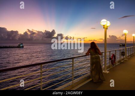 La donna guarda un'oceano da una grande e lussuosa nave da crociera ancorata in un porto durante una torbida e colorato tramonto. Prese a Bridgetown, Barbados. Foto Stock