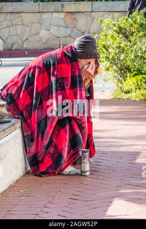 Una persona senza dimora si siede, sporgendoti, su una molto bassa parete dal marciapiede sulla strada statale a Santa Barbara, CA coperto con il rosso e il nero coperta. Foto Stock