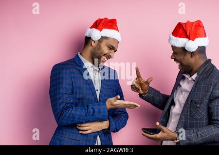 Americano africano e uomo indiano, indossare un abbigliamento formale, in piedi contro la parete di rosa. Due colleghi in cappelli di Babbo Natale, allegramente la comunicazione durante la pausa Foto Stock