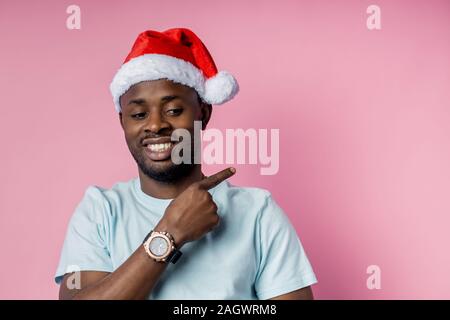 Allegro barba lunga uomo africano indossando Santa hat, bianco di t-shirt, elegante orologio da polso, puntando con il dito indice a lato, mostrando qualcosa di interessante Foto Stock