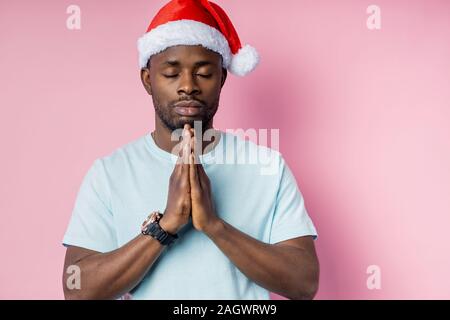 I giovani religiosi calma african american uomo che indossa white t shirt e Santa hat, mantenendo le palme insieme, facendo leva con gli occhi chiusi , sperando che per la migliore, aski Foto Stock