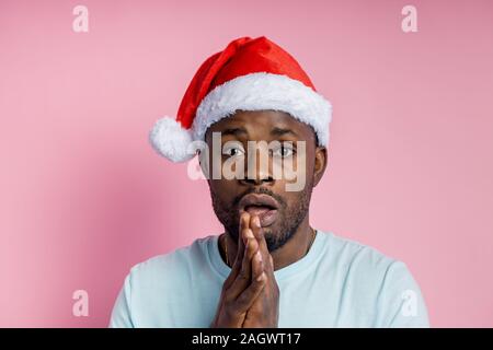 Closeup colpo di scure attraente scuoiati uomo che indossa Santa hat guarda con surprisement, vede qualcosa di stupefacente e mantiene le palme sulle guance, in piedi contro Foto Stock