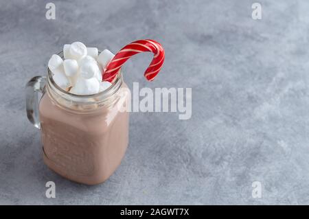 Cioccolata calda con latte, marshmallows e una canna da zucchero in una tazza di vetro su uno sfondo grigio. Anno nuovo scheda con copia spazio. Foto Stock