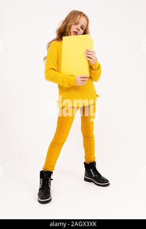 Felice adolescente ragazza con i capelli rossi, felpa con cappuccio e pantaloni gialli letto grande libro giallo isolato su sfondo bianco Foto Stock