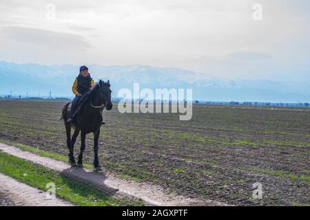 Bishkek, Kirghizistan - 31 Marzo, 2018: bambino uomo a cavallo sul campo di fattoria di montagne vicino al suo villaggio della Repubblica del Kirghizistan Foto Stock