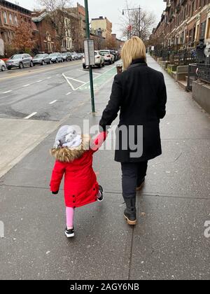 Piccola ragazza di compiere passi da gigante come lei cammina con mom su strada in pendenza del Parco di quartiere, Brooklyn, New York. Foto Stock