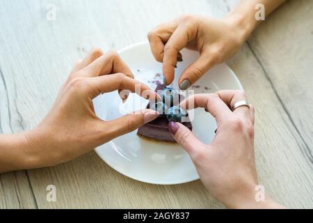 Le mani di due ragazze prendendo i mirtilli dalla parte superiore della gustosa cheesecake su piastra mentre mangiare una torta insieme in cafe Foto Stock