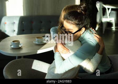 Due giovani affettuosa femminile interculturale dando abbraccio a ogni altra in cafe Foto Stock