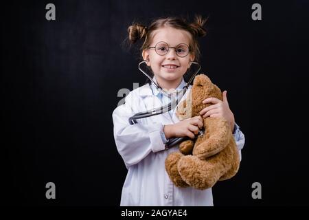 Carino piccolo felice ragazza con uno stetoscopio esaminando orsacchiotto nella parte anteriore della fotocamera Foto Stock