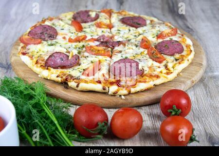 Pane appena sfornato la pizza con salumi, funghi, erbe e un vassoio di formaggi su un tavolo di legno nei pressi di pomodori ciliegini Foto Stock