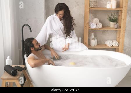Felice giovane donna in accappatoio a parlare con suo marito godendo di vasca da bagno con schiuma Foto Stock