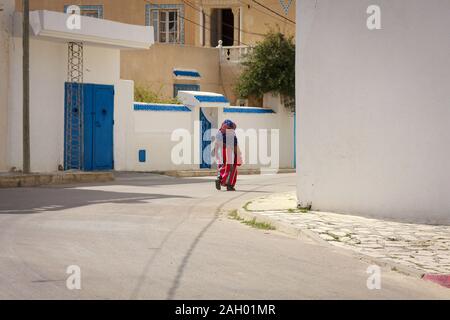 Una donna che cammina per una strada vuota della città tunisina nel bel mezzo della giornata. Edifici con pareti bianche e porte blu e lunghe ombre dal sole di mezzogiorno. Foto Stock