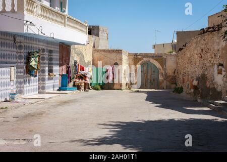 Strada vuota della città tunisina nel mezzo della giornata. Edifici con pareti bianche e porte blu e lunghe ombre dal sole di mezzogiorno, Hergla, Tunisia Foto Stock