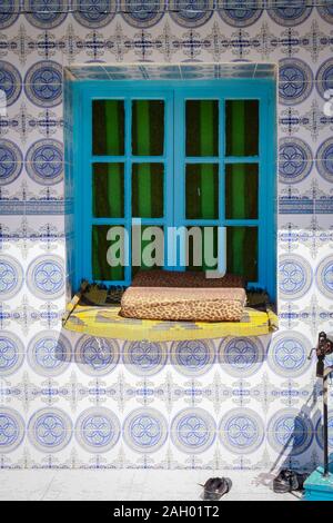 Finestra blu con tende verdi in una parete piastrellata blu della casa con un letto di gatto su un davanzale a Hergla, Tunisia Foto Stock