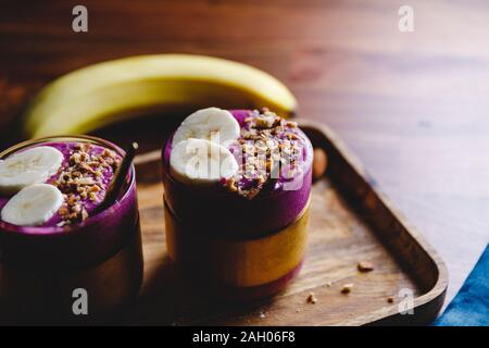 Due bicchieri con violet berry e frullato di banane sono serviti di mandorle schiacciate su un vassoio in legno Foto Stock