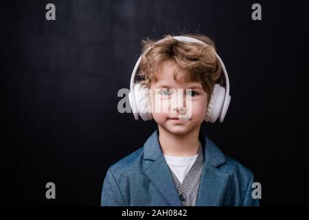 Carino ragazzino informale per ascoltare musica in cuffie bianche nella parte anteriore della fotocamera su sfondo nero Foto Stock