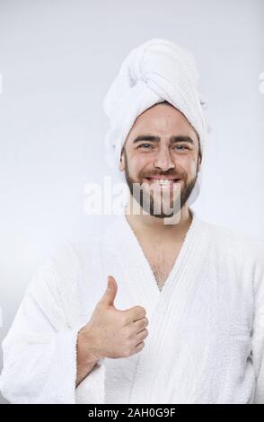 Cintura verticale fino studio Ritratto di Allegro giovane uomo che indossa headwrap asciugamano e accappatoio in piedi con il pollice in su guardando sorridente della fotocamera Foto Stock