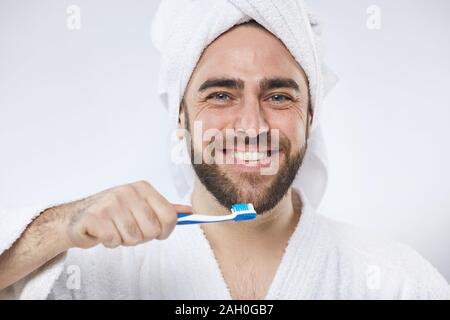 Studio orizzontale ritratto di felice giovane uomo che indossa asciugamano bianco headwrap e accappatoio tenendo lo spazzolino guardando la fotocamera Foto Stock
