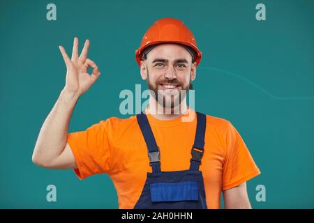 Medium orizzontale ritratto di soggetti di razza caucasica lavoratore edile indossando arancione e blu uniforme che mostra l'anello gesto guardando la telecamera sorridendo, blu sfondo turchese Foto Stock