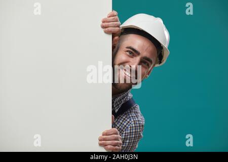 Medium orizzontale immagine ravvicinata di felice lavoratore edile indossando white hardhat fusione da dietro il tabellone vuoto sorridendo, spazio di copia Foto Stock