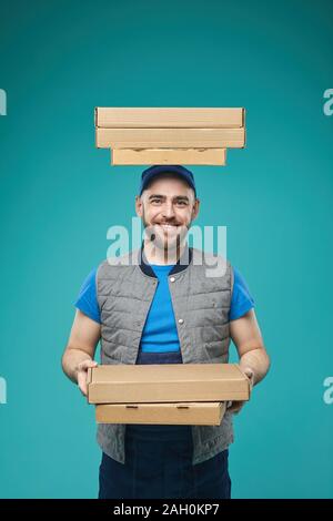 Studio verticale ritratto di consegna del cibo ragazzo con tre scatole per pizza sulla testa portante un altro tre caselle in mani guardando sorridente della fotocamera Foto Stock