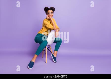 Foto completa del corpo incredibile di pelle scura signora seduta accogliente su una sedia tra le gambe cercando di positivo su telecamera specifiche di usura maglietta gialla pantaloni isolato Foto Stock