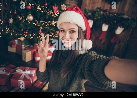 Close up foto di positivo newyear blogger bruna Ragazza capelli in maglia pullover prendere selfie rendere v-segni sul Natale albero sempreverde con molti dono Foto Stock