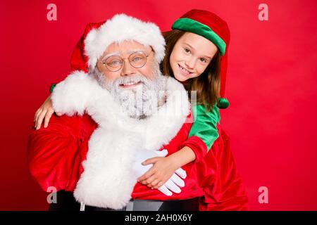 Close up foto di affascinante padre di Natale in occhiali eyewear e il suo elf nel cappello verde cap abbracciando piggyback isolate su sfondo rosso Foto Stock