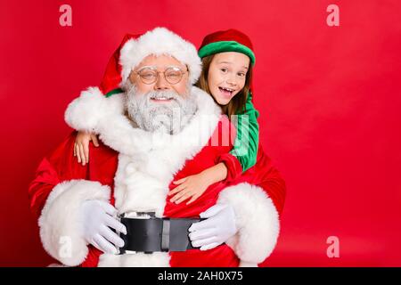 Close up foto di affascinanti santa claus in occhiali eyewear e il suo piccolo elf helper nel cappello verde headwear abbracciando piggyback isolate su rosso Foto Stock