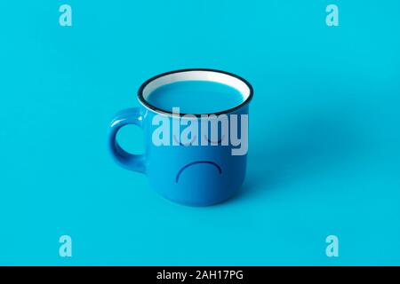 Primo piano di una tazza blu, con un volto triste e disegnati in esso e pieno di un liquido di colore blu su sfondo blu Foto Stock