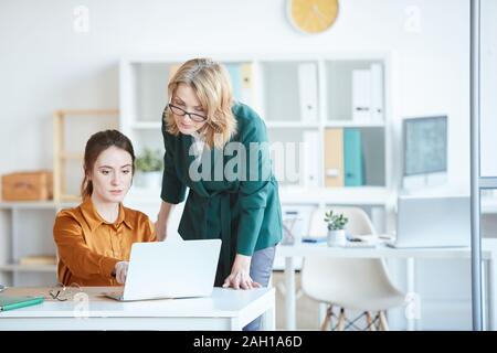 Giovane imprenditrice seduta al tavolo e puntando al monitor di un computer portatile e consultato con il suo collega in ufficio Foto Stock